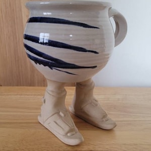 May include: A ceramic mug with a rounded body and a single handle, featuring a cream-colored glaze with horizontal dark blue brushstroke accents. The mug is supported by two sculpted ceramic legs and feet wearing detailed footwear.