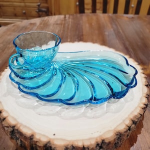 Puede incluir: Un plato de servir de vidrio azul con forma de concha marina con una taza de vidrio azul a juego. El plato y la taza están sobre una rodaja de madera.