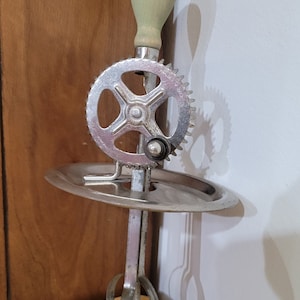 May include: A vintage hand-crank egg beater with a green wooden handle and a silver metal base. The beater has a round, gear-like mechanism and a circular base.