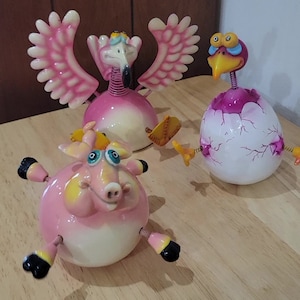 以下が含まれることがあります： それぞれ異なる動物のデザインが施された、風変わりな置物3つ。翼を広げたピンクのフラミンゴ、黄色のアクセントがついたピンクの豚、割れた卵の殻と紫色のアクセントがついた白い鳥。