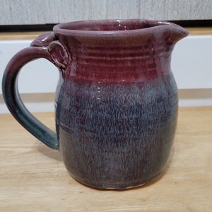 Puede incluir: Una jarra de cerámica hecha a mano con un esmalte morado y azul. La jarra tiene un asa con un detalle decorativo y un pico.