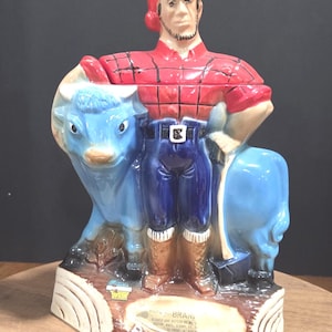 Peut inclure: Figurine en céramique de Paul Bunyan avec un bœuf bleu. La figurine porte une chemise à carreaux rouge, un pantalon bleu et un bonnet rouge. La base est marron avec le nom "Paul Bunyan" en lettres blanches.