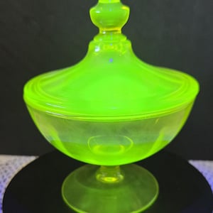 May include: A vibrant neon yellow, translucent glass candy dish with a matching lid. The dish features a wide, rounded bowl shape supported by a circular pedestal base, topped with a pointed finial handle on the lid, glowing under UV light.