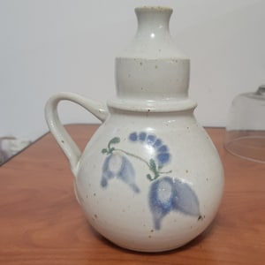 Puede incluir: Una jarra de cerámica blanca con un diseño floral azul y verde. La jarra tiene un cuerpo redondo y un pico. Tiene una asa en el lateral.