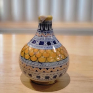 Puede incluir: Un pequeño jarrón redondo de cerámica con un patrón geométrico amarillo, azul y blanco. El jarrón tiene un cuello estrecho y una base ancha.