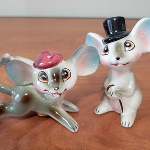 Puede incluir: Dos figuritas de ratón de cerámica. Un ratón lleva una boina roja y el otro un sombrero de copa negro. Ambos ratones tienen detalles pintados y orejas grandes.