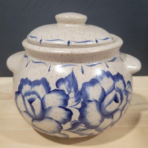 Pode incluir: Uma panela de cerâmica branca com tampa, decorada com desenhos florais azuis. A panela tem duas alças nas laterais e um esmalte salpicado.