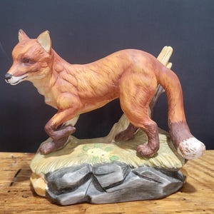 Puede incluir: Figura de zorro de cerámica en tonos naranja, marrón y blanco. El zorro está posado sobre una base rocosa con hierba y pequeñas flores. La figura tiene detalles y es un artículo decorativo.