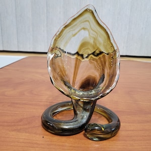 May include: A green glass vase shaped like a snake with a flared opening. The vase has a swirl pattern and is made of clear glass with a green tint.