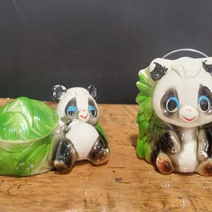 Puede incluir: Dos figuras de oso panda de cerámica. Un panda sostiene un recipiente en forma de col verde. El otro panda tiene hojas verdes a su alrededor. Ambos pandas tienen pelaje blanco y negro, ojos azules y sonrisas rojas.