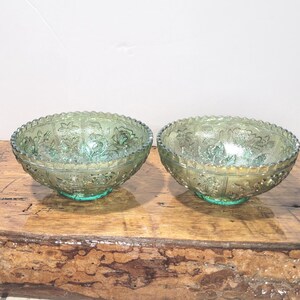 May include: Two matching green glass bowls with floral patterns. The bowls have a scalloped rim and sit on a green base. The bowls are on a wooden surface.