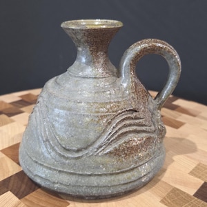 Pode incluir: Jarro de cerâmica feito à mão com uma alça. O jarro é principalmente cinza com detalhes em marrom e uma superfície texturizada. O design inclui padrões ondulados esculpidos na lateral e uma borda alargada. O jarro está em uma tábua de corte de madeira.