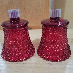Könnte beinhalten: Zwei rote Glas-Kerzenhalter mit einem strukturierten, blasenartigen Muster. Jeder Halter hat einen ausgestellten Sockel, eine abgerundete Oberseite und einen transparenten Kunststoffeinsatz. Die Kerzenhalter werden auf einer hellen Holzoberfläche präsentiert.