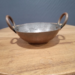 Peut inclure: Un petit bol en cuivre martelé avec deux anses. Le bol est posé sur une surface en bois.