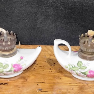 Puede incluir: Dos lámparas de aceite antiguas de porcelana blanca con diseños florales. Cada lámpara tiene un asa, un quemador de latón y una mecha. Las lámparas están decoradas con rosas rosas y hojas verdes, y se exhiben sobre una superficie de madera.