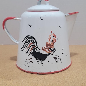 Könnte beinhalten: Weiße Emaille-Kaffeekanne mit rotem Rand, Griff und Deckelakzent. Die Kanne zeigt eine Hahn-Illustration in Schwarz, Braun und Rot. Der Ausguss ist abgewinkelt. Ein Vintage-Küchenartikel.