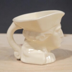 Può includere: Una tazza in ceramica bianca a forma di testa di persona con un cappello. La tazza ha un manico ed è progettata per contenere una piccola quantità di liquido.