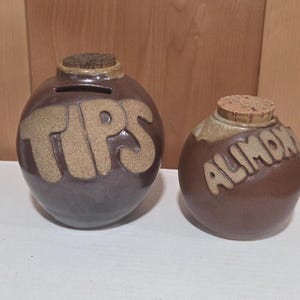 May include: Two round, brown ceramic containers with cork stoppers. The larger container features a horizontal slot and the word TIPS in raised, textured lettering. The smaller container features the word ALIMONY in raised, textured lettering.