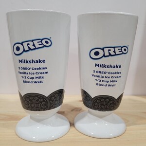 May include: Two white milkshake glasses with the Oreo logo and milkshake recipe printed on them. The glasses have a wide base and a tall, slightly tapered body. The bottom of the glasses feature a black and white Oreo cookie design.