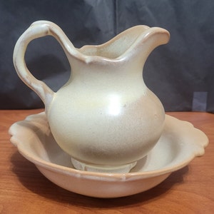 May include: A cream-colored ceramic creamer and sugar bowl set with a textured glaze. The creamer has a handle and a rounded body. The sugar bowl is round and has a scalloped edge.
