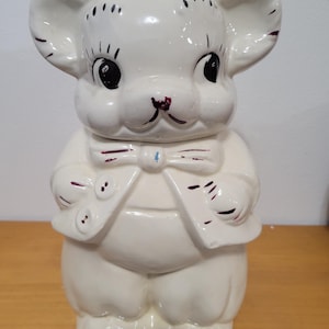 Puede incluir: Tarro de galletas de cerámica blanca con forma de oso que lleva una pajarita burdeos y una chaqueta burdeos con botones blancos. El oso tiene ojos negros y una nariz burdeos.