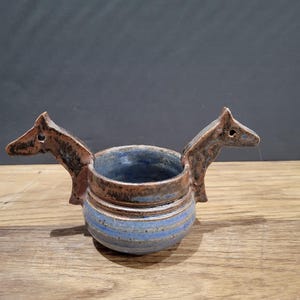 May include: A small, blue and brown ceramic pot with two handles shaped like horse heads.