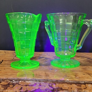 Könnte beinhalten: Zwei Dessertschalen aus leuchtend grünem Glas im Vintage-Stil. Die Schalen haben ein Gittermuster und stehen auf einem runden Sockel. Eine Schale hat einen Griff. Das Glas hat einen leichten Glanz. Der Hintergrund ist dunkel.
