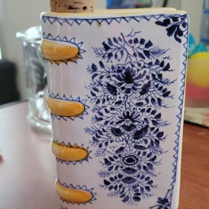 Może przedstawiać: Ceramiczna karafka w kształcie książki z korkowym zatyczką. Biała powierzchnia posiada niebieski wzór kwiatowy oraz żółte, wypukłe poziome pasy na grzbiecie. Krawędzie są ozdobione niebieską bordiurą w zygzaki oraz niebieskimi motywami kwiatowymi.