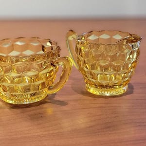 Puede incluir: Un par de piezas de servicio de vidrio ámbar vintage, una jarra de crema y un azucarero. Ambas piezas tienen un patrón de diamantes y asas. La jarra de crema tiene un pico. El juego está sobre una superficie de madera.