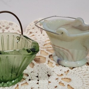 May include: Two small glass ashtrays shaped like buckets. One is green glass with a brass handle and the other is white with swirls of purple and brown.