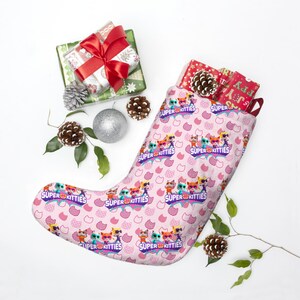 May include: A pink Christmas stocking with a repeating pattern of cartoon cats and the words "SUPER KITTIES." The stocking is filled with wrapped gifts and pine cones. A wrapped gift with a red bow and a silver ornament are also present.
