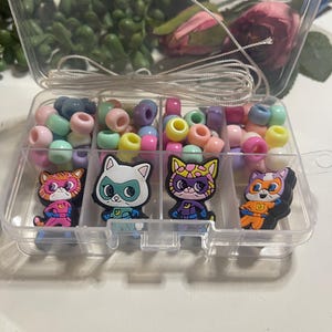 May include: A clear plastic container with six compartments filled with colourful beads and four small character figurines. The figurines are of cats wearing superhero costumes.