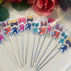 May include: A selection of cat-themed cupcake toppers. Each topper depicts a cartoon cat in a superhero costume and mask. The cats are in various colours, including blue, orange, pink, and yellow. The toppers are attached to clear plastic sticks.