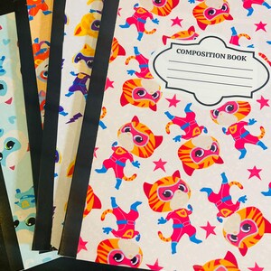 May include: A stack of composition notebooks with black spines. The notebooks feature colorful covers with cartoon superhero cat illustrations in red, orange, and blue. The text "COMPOSITION BOOK" is visible on one cover.