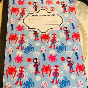 May include: A composition book with a light blue cover featuring a pattern of red, blue, and white Spider-Man characters, spiders, and spiderwebs. The cover includes the text "COMPOSITION BOOK" within a white label.