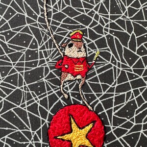 May include: Embroidered image of a small animal in a red circus outfit, balancing on a red ball with a gold star. The background is black with a white web-like pattern. The animal is holding a gold baton.