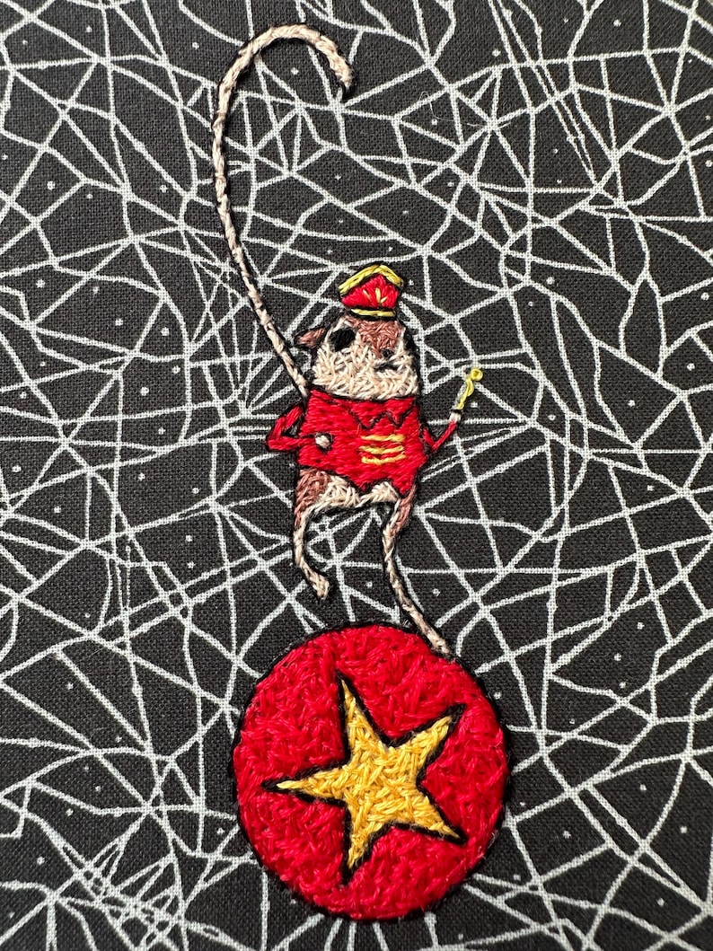 May include: Embroidered image of a small animal in a red circus outfit, balancing on a red ball with a gold star. The background is black with a white web-like pattern. The animal is holding a gold baton.