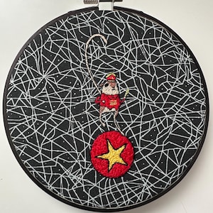 May include: Embroidered hoop art featuring a circus mouse balancing on a red ball with a gold star. The mouse wears a red coat and hat. The background is black with a white web-like pattern. The hoop is black.