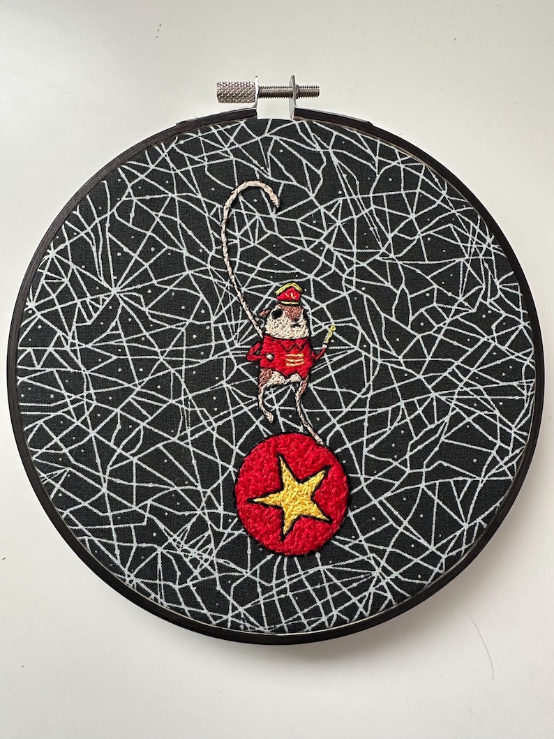 May include: Embroidered hoop art featuring a circus mouse balancing on a red ball with a gold star. The mouse wears a red coat and hat. The background is black with a white web-like pattern. The hoop is black.