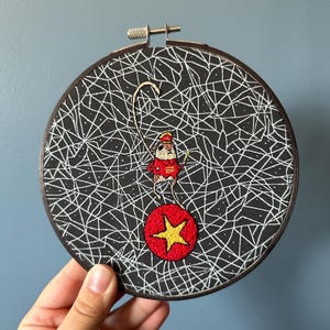 May include: Embroidered artwork in a round wooden hoop. The black fabric background features a white web-like design. A small figure in a red outfit balances on a red ball with a yellow star. The hoop is held by a hand.