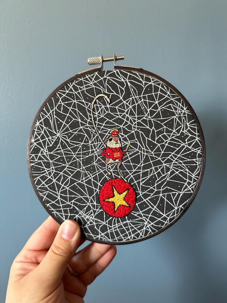 May include: Embroidered artwork in a round wooden hoop. The black fabric background features a white web-like design. A small figure in a red outfit balances on a red ball with a yellow star. The hoop is held by a hand.