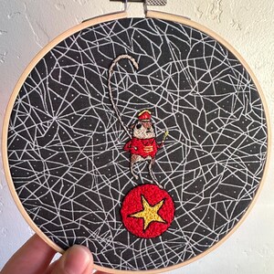 May include: Embroidered hoop art featuring a circus mouse balancing on a red ball with a gold star. The mouse wears a red jacket and hat, holding a whip. The background is black with a white web-like pattern.