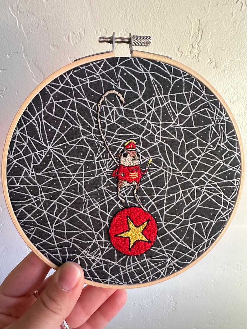 May include: Embroidered hoop art featuring a circus mouse balancing on a red ball with a gold star. The mouse wears a red jacket and hat, holding a whip. The background is black with a white web-like pattern.