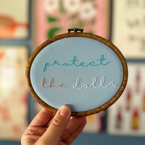 May include: A round embroidery hoop with a light blue fabric background. The words "protect the dolls" are embroidered in teal, pink, and white thread. The hoop has a dark wooden frame.