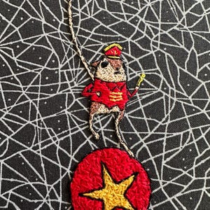 May include: Embroidered circus-themed artwork featuring a mouse in a red and gold uniform, balancing on a red ball with a gold star. The background is black with a white web-like pattern. The mouse holds a whip.