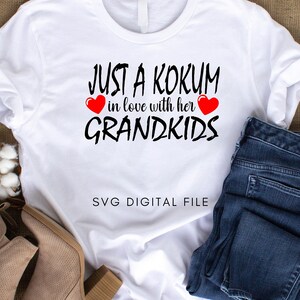 Può includere: Maglietta bianca con testo nero che recita "Just a Kokum in love with her grandkids" con due cuori rossi. Il testo è in stile calligrafico. La maglietta è su uno sfondo di juta marrone con un paio di stivaletti marroni alla caviglia e un paio di jeans blu.