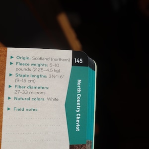 Peut inclure: Gros plan sur une page de livre avec des informations sur le mouton North Country Cheviot. Le texte comprend l'origine, le poids de la toison (2,25-4,5 kg), la longueur des fibres (9-15 cm), le diam&egrave;tre des fibres et les couleurs naturelles. Le num&eacute;ro de page 145 est visible.