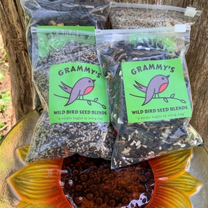 May include: Three bags of wild bird seed blends with a robin logo and the text "GRAMMY'S WILD BIRD SEED BLENDS" and "a birdie buffet in every bag". The bags are sitting on a yellow and orange sunflower-shaped bird feeder.