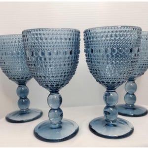 Może przedstawiać: Cztery niebieskie kieliszki do wina z teksturowanym wzorem przypominającym bąbelki. Każdy kieliszek ma okrągłą podstawę, nóżkę z dwoma sferycznymi akcentami i szeroką czarkę. Kieliszki są ustawione na białej powierzchni.