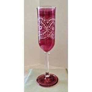 May include: A tall, ruby-colored champagne flute with a clear stem and base. The glass is decorated with a white, repeating floral pattern around the upper portion. The flute is displayed on a light green surface.